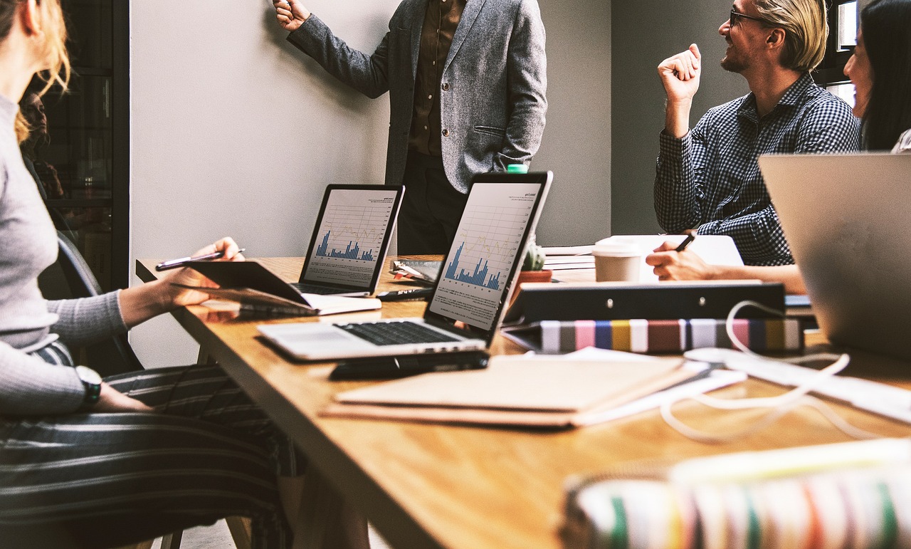 Personen arbeiten an Laptops im Büro, während eine Person steht und etwas präsentiert.